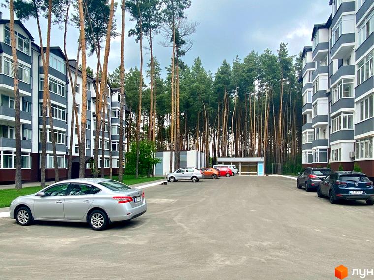 Житловий комплекс Ідея з сіро-білими фасадами, оточений високими соснами та парковкою.