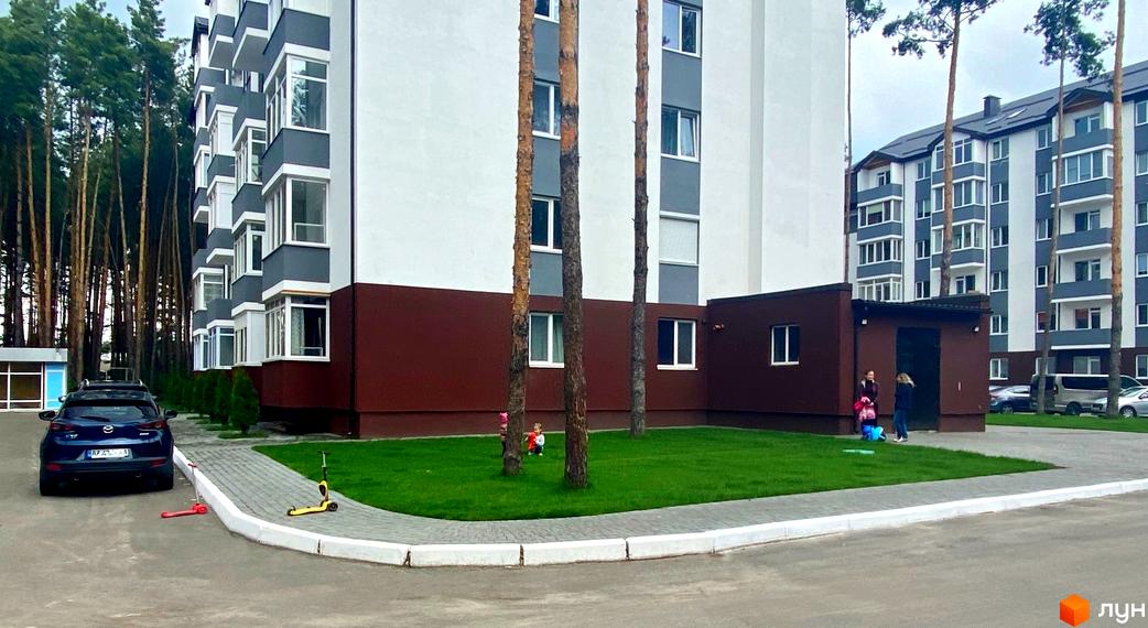 Житловий комплекс Ідея з біло-сірими фасадами та балконами. Прибудинкова територія має доглянутий газон, сосни та дитячі самокати.