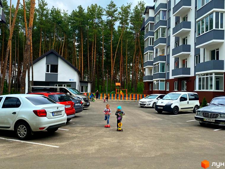 Житловий комплекс Ідея з біло-сірими фасадами та балконами. Поруч парковка, дитячий майданчик та сосновий ліс.