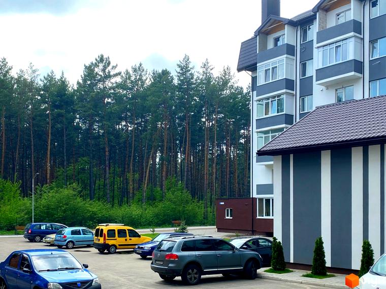Житловий комплекс Ідея: багатоповерховий будинок з сіро-білим фасадом та коричневим дахом. Поруч парковка та сосновий ліс.