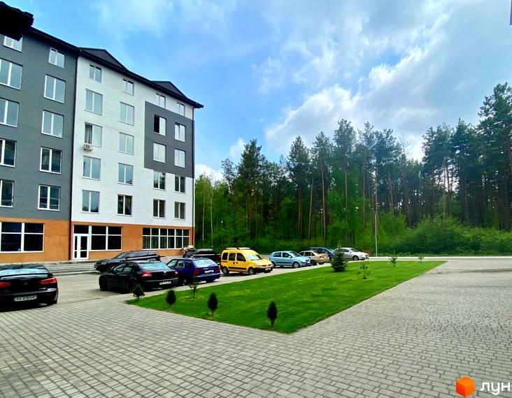 Багатоповерховий будинок Житлового комплексу Ідея з сіро-білим фасадом та великими вікнами. Перед будинком розташований паркінг, зелена галявина та ліс.