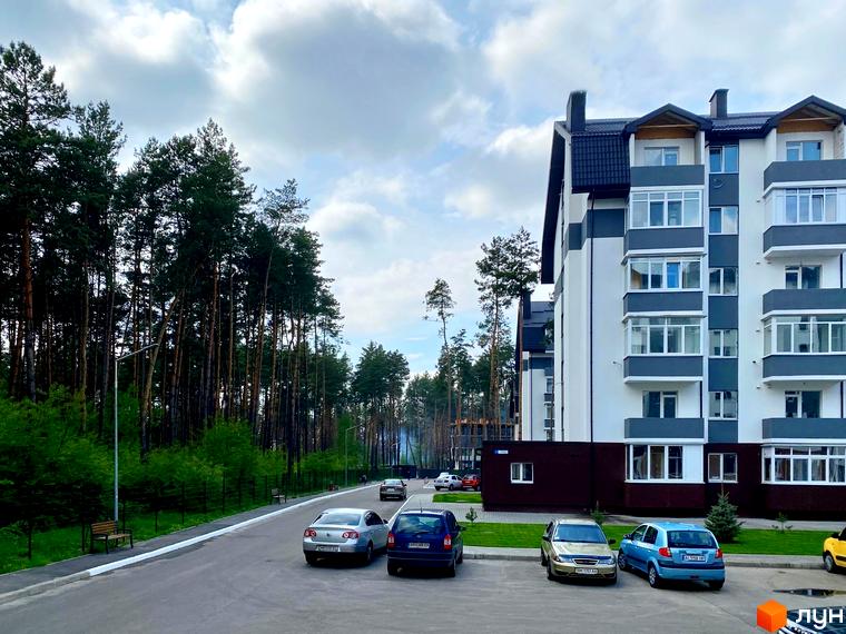 Житловий комплекс Ідея з біло-сірим фасадом та балконами. Поруч дорога, парковка для автомобілів та сосновий ліс.