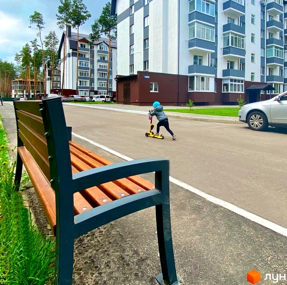 Багатоповерхові будинки Житлового комплексу Ідея з біло-сірими фасадами та балконами. Прибудинкова територія з дорогою, зеленою травою та лавкою.