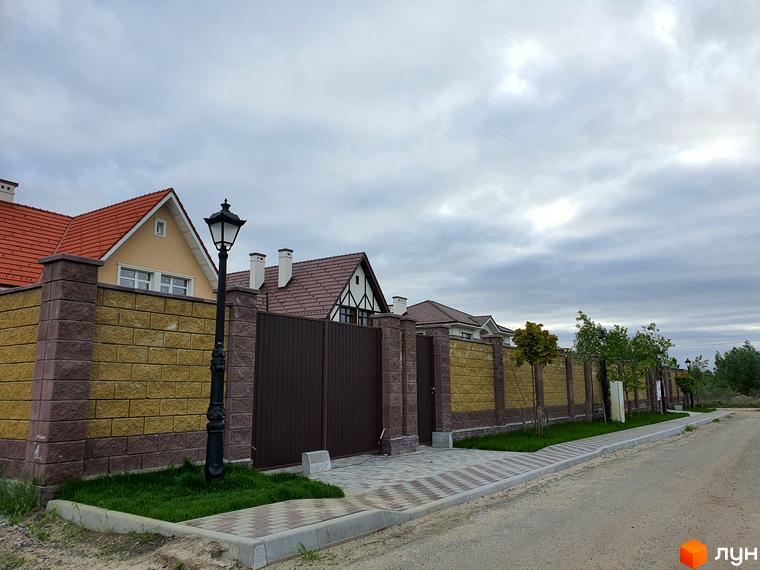Зображення фасаду будинку в Містечку котеджів КМ Осокор. Видно цегляний паркан, ковані ворота та ліхтарі вздовж дороги.