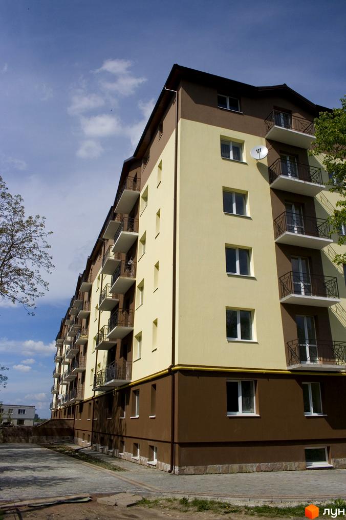 Зовнішній вигляд новобудови ЖК Підгір'я з балконами та вікнами на фоні блакитного неба з хмарами. Прибудинкова територія з бруківкою та зеленими насадженнями.