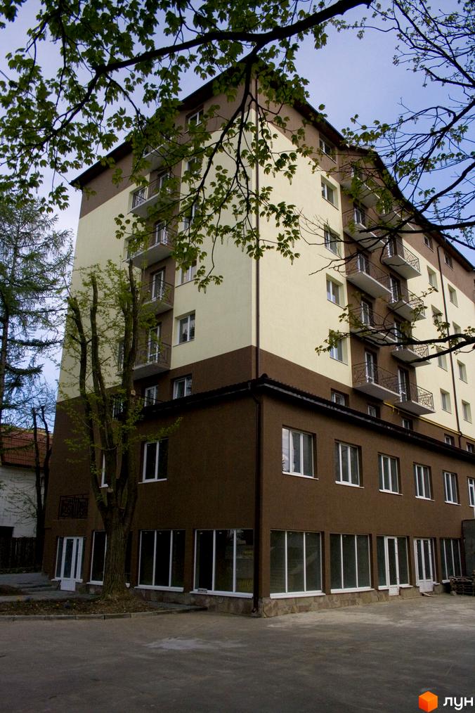 Зовнішній вигляд багатоповерхового будинку ЖК Підгір'я з балконами та вікнами на тлі дерев та блакитного неба. Прибудинкова територія з асфальтованим покриттям та зеленими насадженнями.