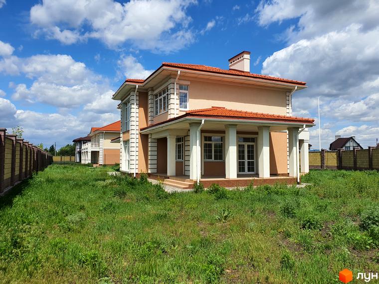 Двоповерховий будинок з червоною черепицею на даху та білими колонами на терасі. Перед будинком зелена трава та газон, а також паркан.