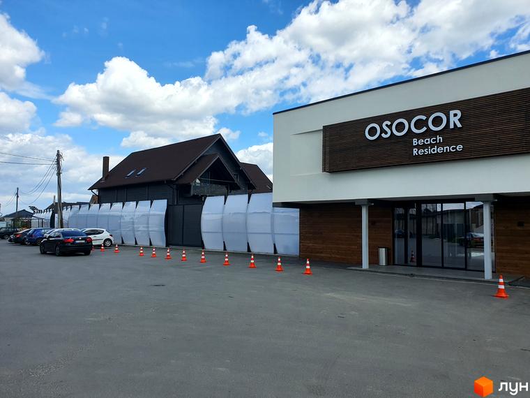 Фасад будівлі Комплекс Осокор Beach Residence з великими вікнами та вивіскою. Перед будівлею парковка та дорожні конуси, на задньому плані котеджі та блакитне небо.