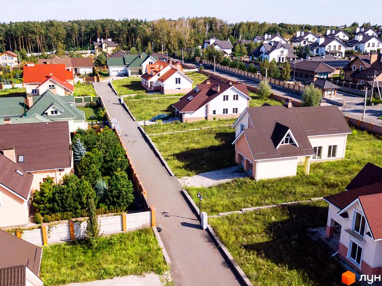 Котеджне містечко На узліссі: двоповерхові будинки з коричневими дахами та світлими фасадами. Навколо будинків – зелені газони та дерева, асфальтована дорога.