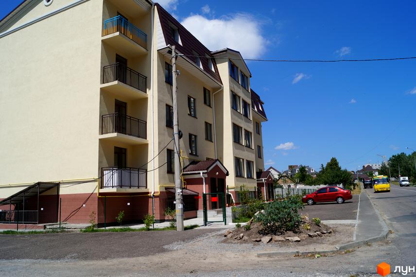 Багатоповерховий Клубний будинок Приозерний 24 з бежевим фасадом, балконами та коричневим дахом. Поруч дорога, прибудинкова територія з огорожею та автомобілями.