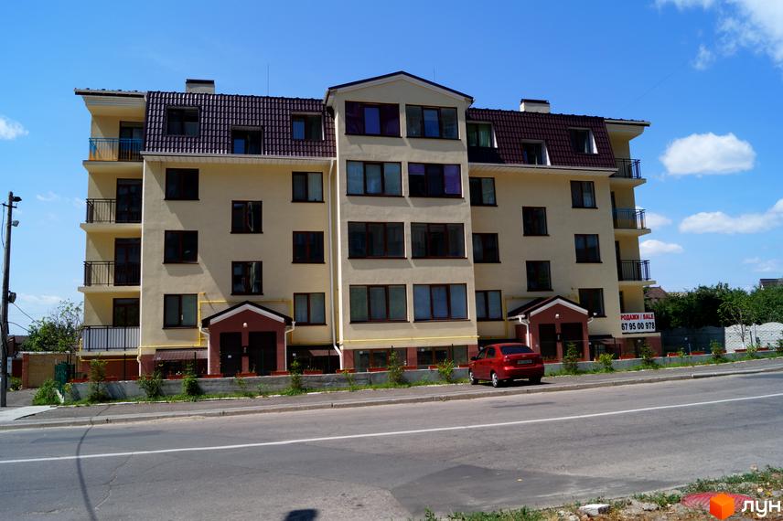 Клубний будинок „Приозерний 24“: бежева багатоповерхова будівля з коричневим дахом та балконами. Перед будинком дорога, зелені насадження та припаркований автомобіль.