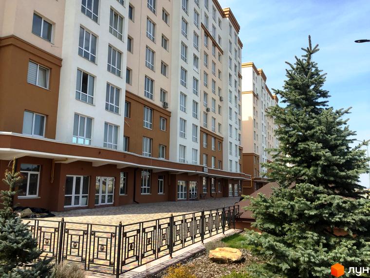 Житловий комплекс Львівський маєток. Багатоповерхові будинки з біло-коричневим фасадом та великими вікнами. Поруч зелена ялина та огороджена територія.