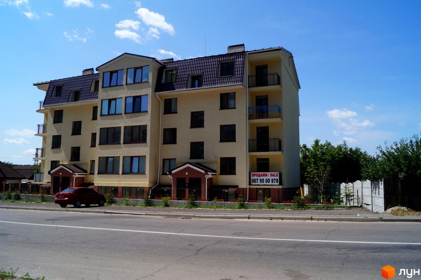 Багатоповерховий клубний будинок Приозерний 24 світло-жовтого кольору з темним дахом та балконами. Поруч дорога, автомобіль та зелені насадження.