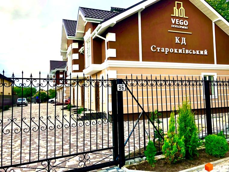 Таунхауси «КД Старокиївський»: фасади будинків з коричневими стінами та білими вікнами, огороджені чорним металевим парканом.