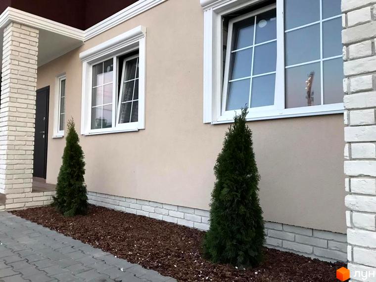 Зовнішній вигляд будинку з білою цегляною обробкою та вікнами, що виходять на зелену прибудинкову територію з деревами.