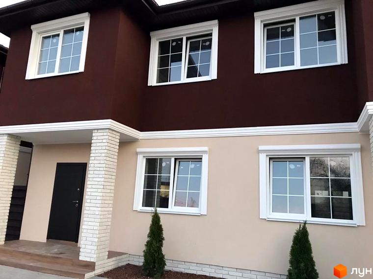 Фасад двоповерхового будинку з білими вікнами та оздобленням. Біля входу колони з білої цегли, на території зелені насадження.