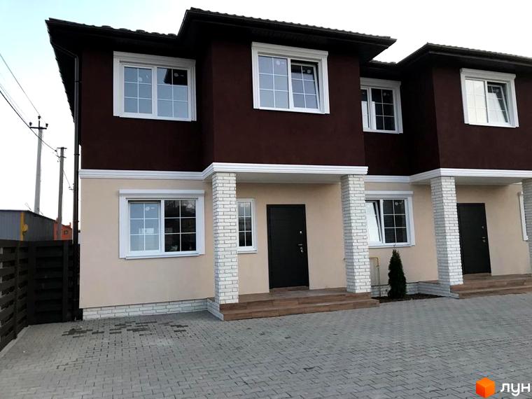 Фасад будинку житлового комплексу з коричневою та бежевою штукатуркою, білими вікнами та колонами, бруківка перед будинком.