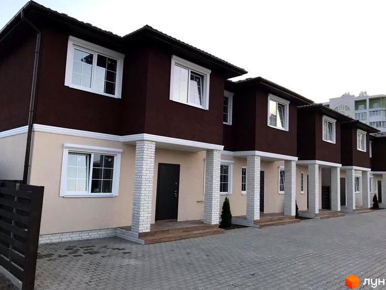 Зовнішній вигляд будинку житлового комплексу з коричневим фасадом та білими вікнами. Бруківка на прибудинковій території.