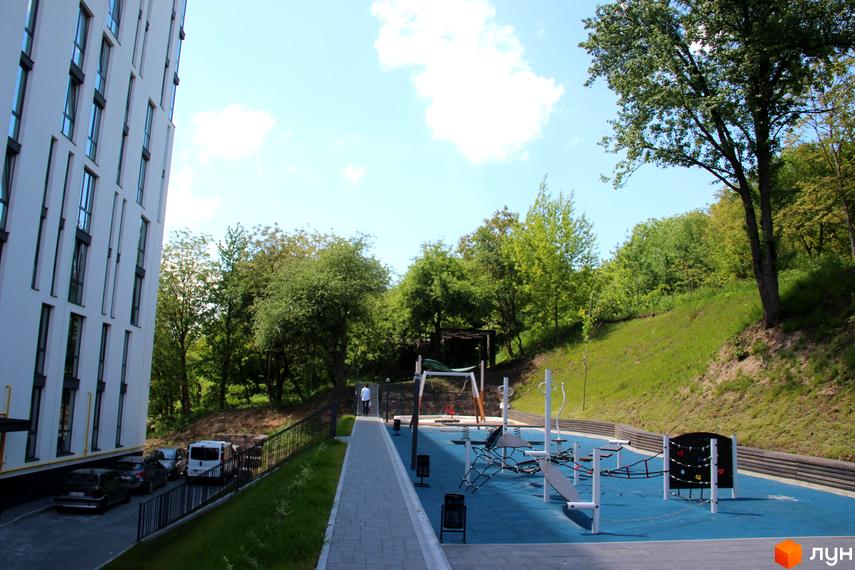 Новобудова ЖК OBRIY з білим фасадом та панорамними вікнами. Поруч знаходиться дитячий майданчик з різноманітним обладнанням та зелена зона.