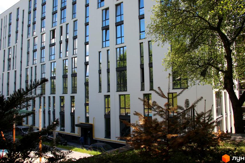 Фасад житлового комплексу OBRIY з великою кількістю вікон та зеленою прибудинковою територією. Будинок має білий колір та темні віконні рами.