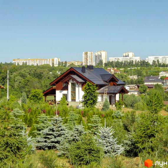 Котеджне містечко Шалє Грааль: двоповерховий будинок з темним дахом та білими стінами, оточений зеленню та деревами. На задньому плані видніються багатоповерхові будівлі.