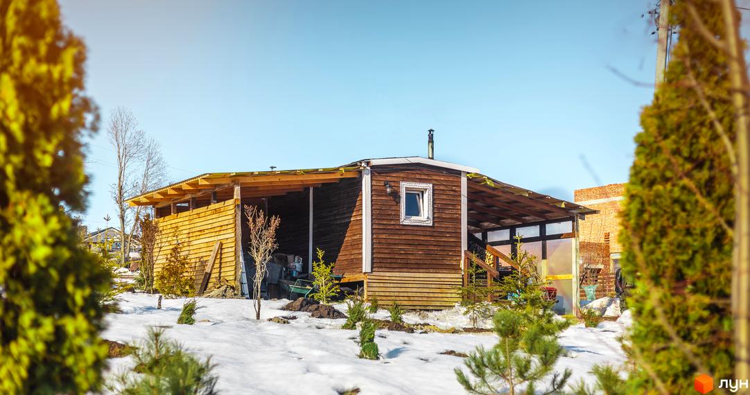 Дерев'яний будинок з терасою та вікном, розташований серед засніженого ландшафту. Навколо будинку ростуть дерева та кущі, створюючи затишну атмосферу.