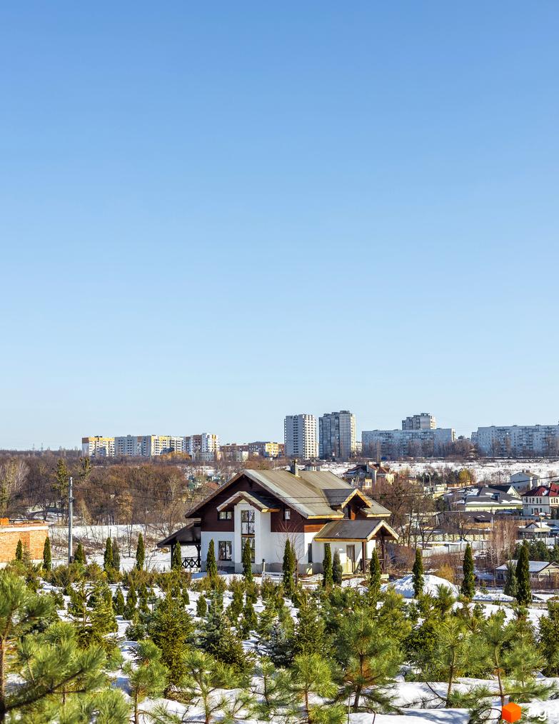 Котеджне містечко Шалє Грааль: двоповерховий будинок з коричневим дахом та білими стінами, оточений зеленню та снігом. На задньому плані видніються багатоповерхові будівлі.