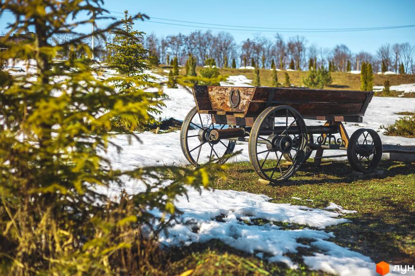 Дерев'яний віз стоїть на тлі засніженого ландшафту. Поруч з возом зелені дерева та кущі, що створюють контраст з зимовим пейзажем.
