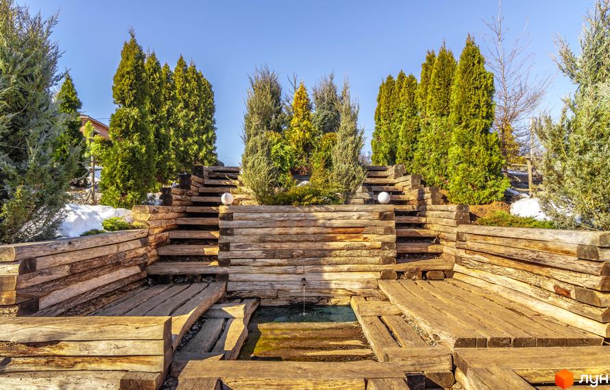 Зовнішній вигляд будинку КМ Шале Грааль з дерев'яними сходами та декоративним водоймищем. Прибудинкова територія з туями та снігом.