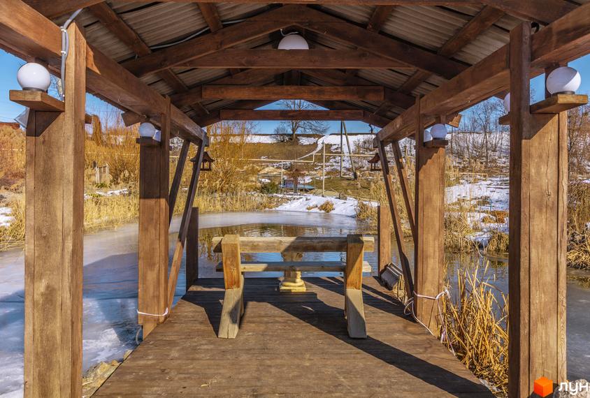 Дерев'яний навіс з лавкою над замерзлою водоймою в Котеджному містечку Шалє Грааль. Навкруги сніг та рослинність.