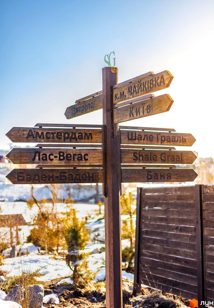 Дерев'яний вказівник з назвами напрямків. Поряд розташована частина дерев'яного паркану та засніжена територія.