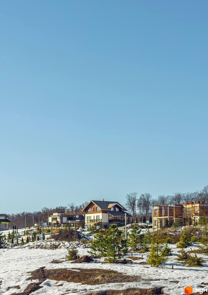 Котеджне містечко Шале Грааль: двоповерхові будинки з коричневими дахами та дерев'яними елементами на тлі блакитного неба. Навколо – ділянка зі снігом та молодими соснами.