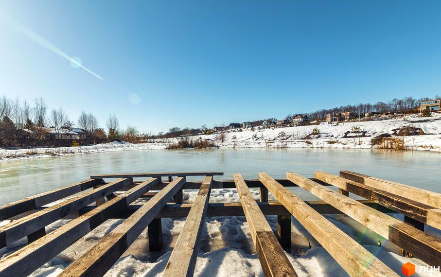 Котеджне містечко Шалє Грааль розташоване на березі замерзлої водойми, з дерев'яним пірсом попереду та будинками на фоні засніженого схилу.