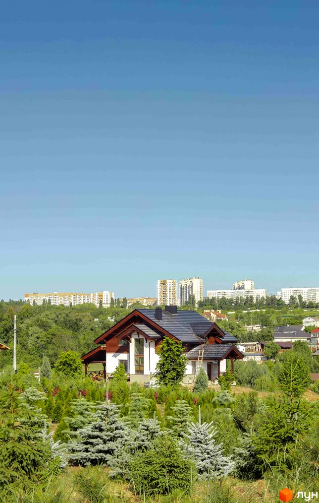 Будинок з темно-коричневим дахом та білими стінами, оточений зеленими деревами та рослинами. На задньому плані видно багатоповерхівки.