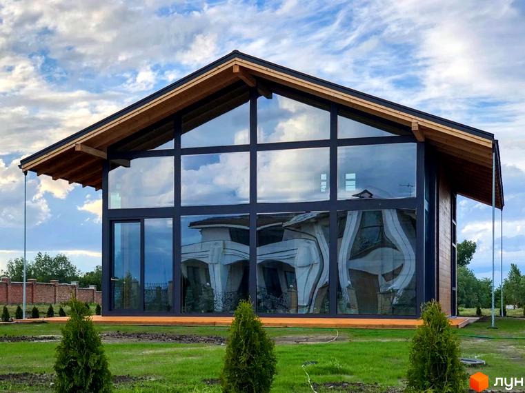 Котеджне містечко КМ Riviera Sunrise з панорамними вікнами та дерев'яними елементами фасаду. Навколо будинку зелена галявина та молоді туї.