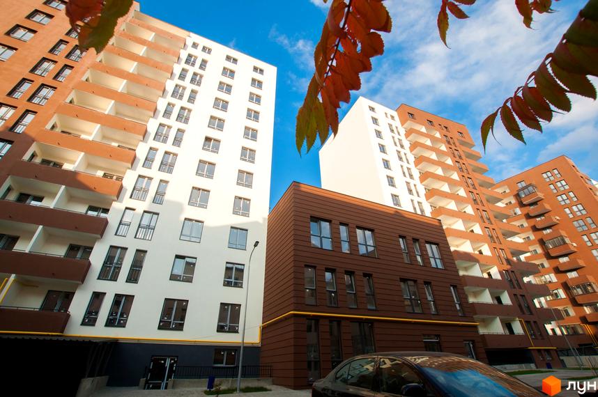 Зовнішній вигляд житлового комплексу Еко-дім на Мечникова 2, з фасадами білого та коричневого кольору, балконами та вікнами. Навколо новобудови видно дерева та блакитне небо.