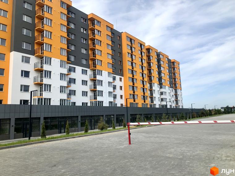 Зовнішній вигляд новобудови ЖК Сімейний COMFORT з помаранчевими та білими фасадами. Біля будинку облаштована прибудинкова територія з парканом та шлагбаумом.