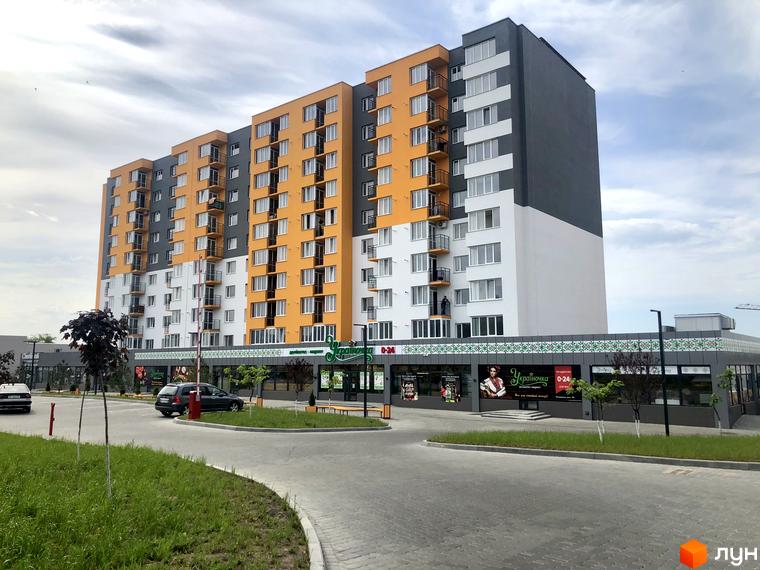 Зовнішній вигляд новобудови ЖК Сімейний COMFORT з яскравою фасадною обробкою та прибудинковою територією з дорогою та зеленню. Комплекс має багатоповерхові секції з балконами та великими вікнами.