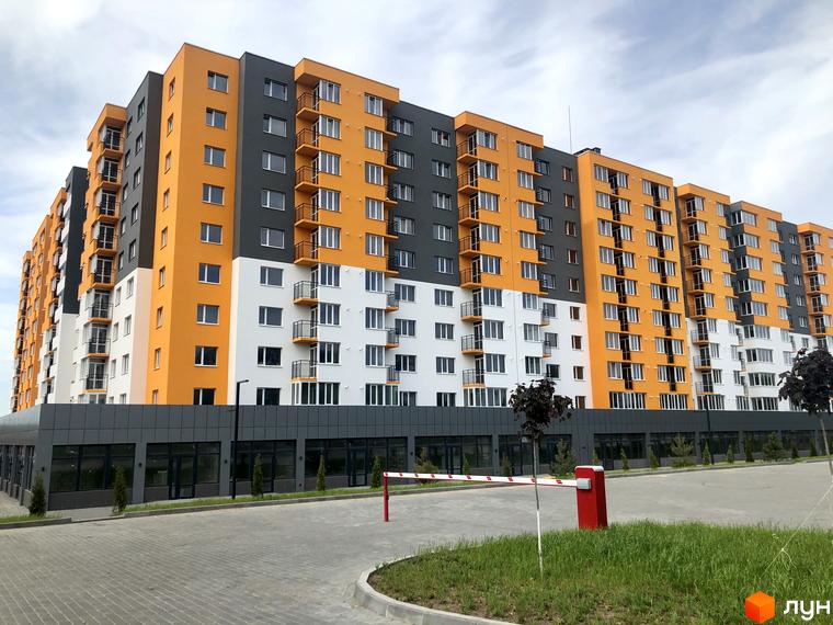 Житловий комплекс Сімейний COMFORT з яскравим фасадом з помаранчевих та сірих секцій. Перед будинком розташована прибудинкова територія з дорогою та шлагбаумом.