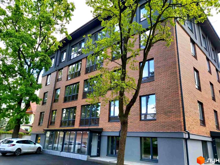 Зовнішній вигляд житлового комплексу Brighton Residence з цегляним фасадом, великими вікнами та зеленню навколо. Біля будинку припаркований білий автомобіль.