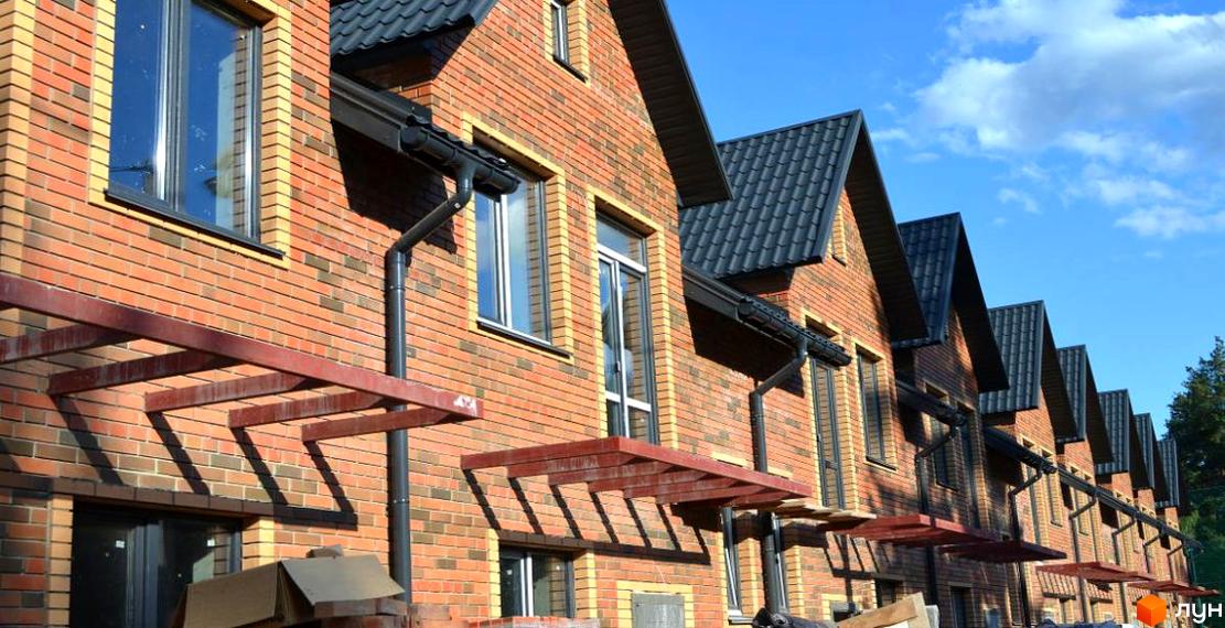 Цегляні будинки з темною покрівлею та вікнами, розташовані в ряд. На фасадах є навіси, а на небі видно блакитне небо з хмарами.