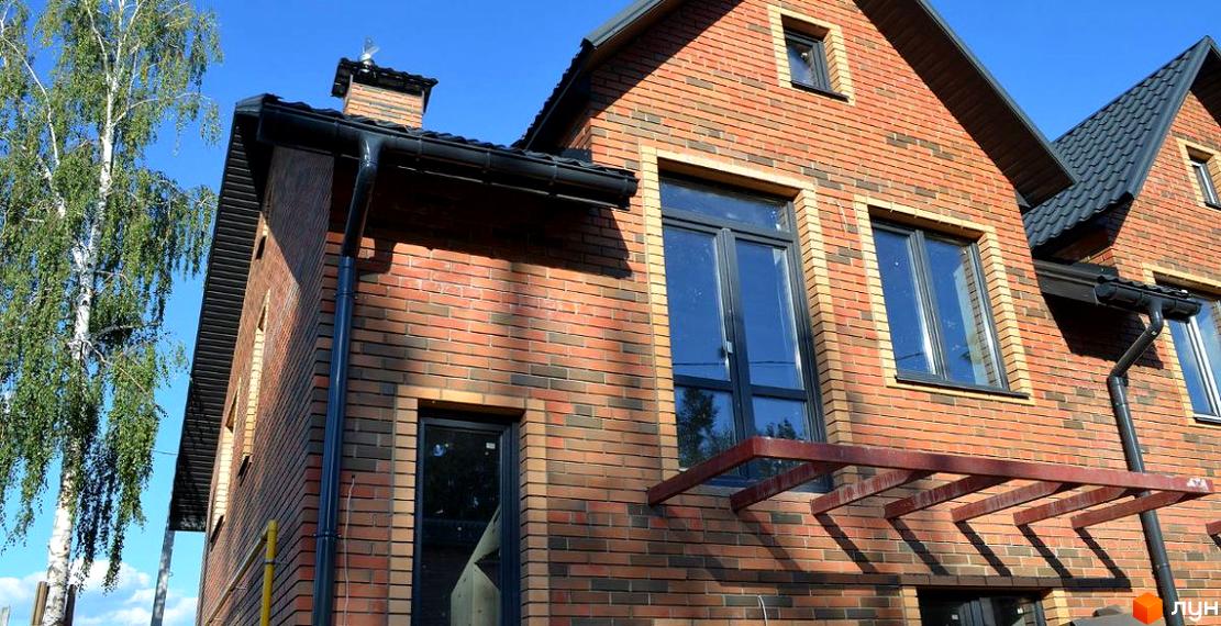 Фасад цегляного будинку з великими вікнами та чорною покрівлею. Навколо будинку видно зелене дерево та блакитне небо.