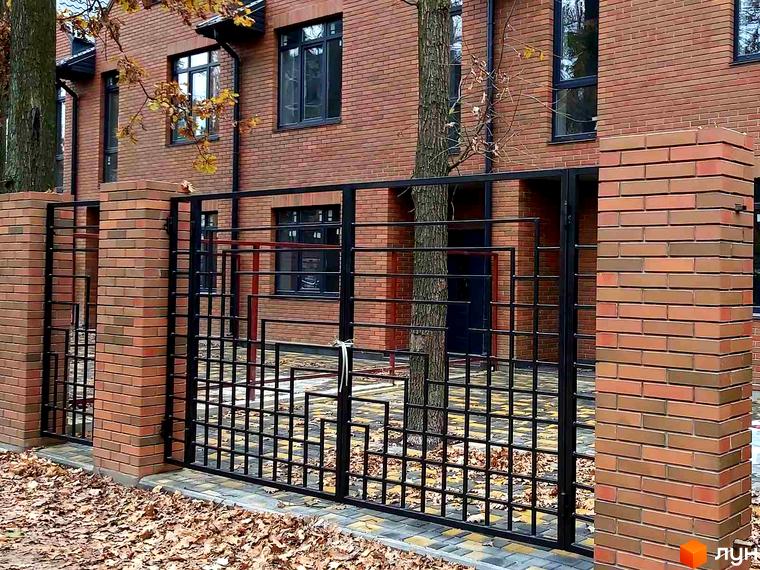 Цегляний будинок з кованим парканом та колонами. На території осіннє листя, росте дерево.
