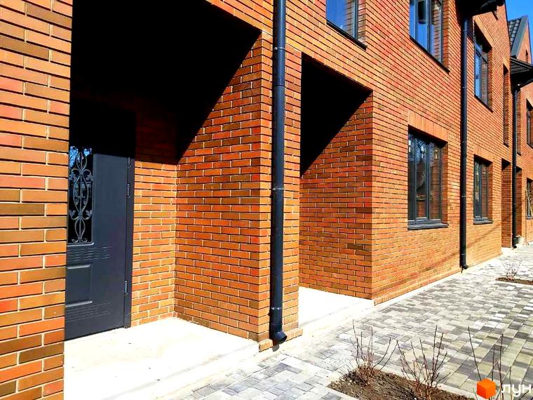 Таунхауси Brighton 3 з червоної цегли з сірими вікнами та дверима. Перед будинками викладена бруківка та висаджені молоді дерева.