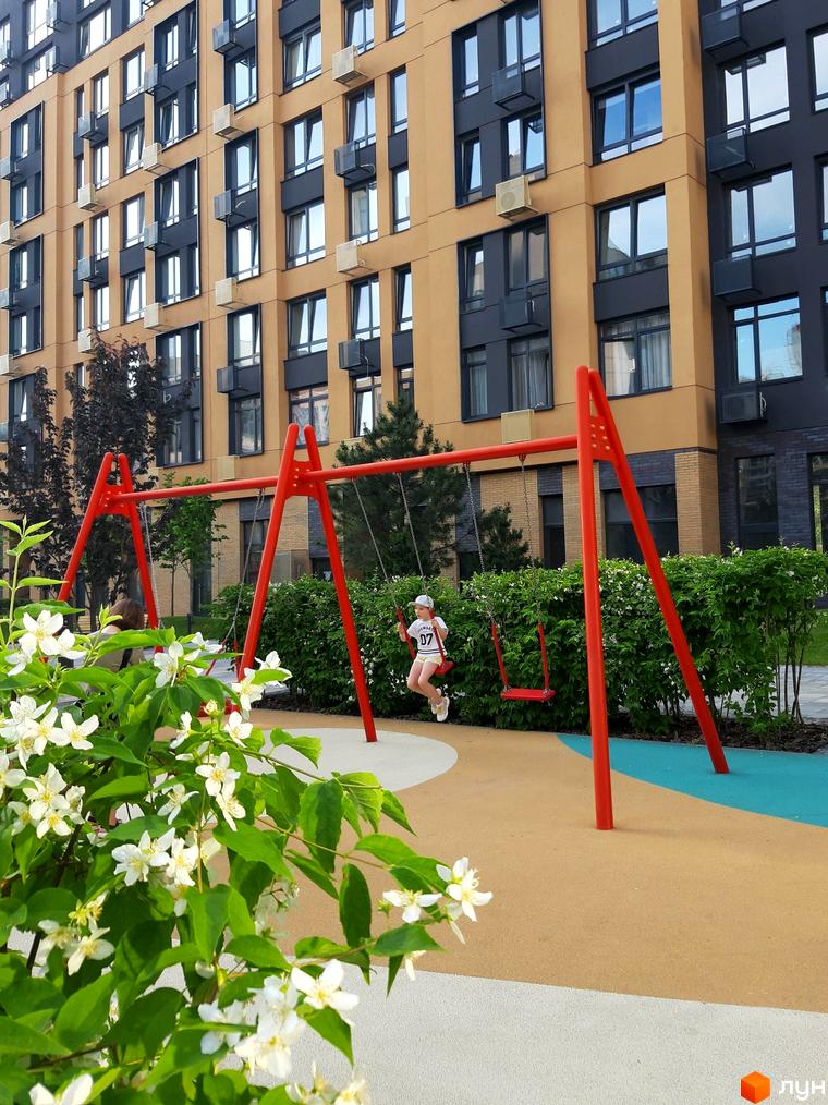 Житловий комплекс Файна Таун з багатоповерховим будинком у світло-коричневих та темно-сірих тонах. На передньому плані дитячий майданчик з червоними гойдалками та квітучими кущами.