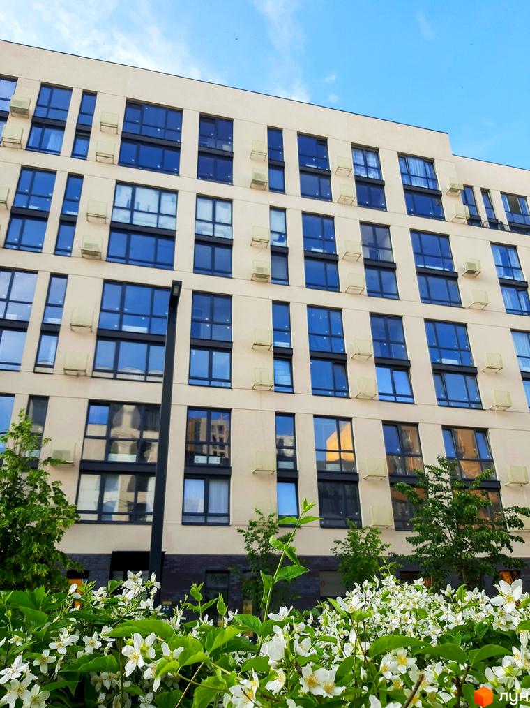 Висотна будівля з великою кількістю вікон та кондиціонерами на фасаді. Перед будинком розташовані квіти та дерева.