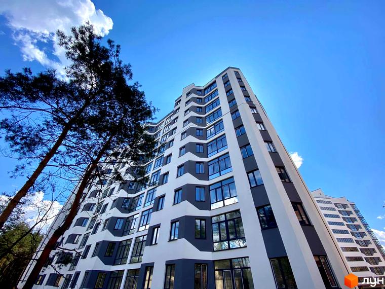 Житловий комплекс Авіатор - сучасна новобудова з панорамними вікнами та незвичайною архітектурою. Будинок оточений деревами під яскравим блакитним небом.