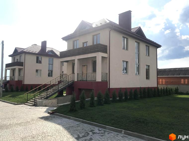 Будинки Котеджного містечка Горобиновий маєток зі світлими фасадами, темними дахами та зовнішніми сходами. Навколо будинків зелений газон та бруківка.