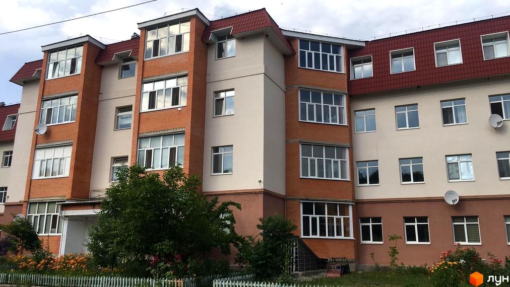 Багатоквартирний будинок з червоною цеглою та світлою штукатуркою, червоним дахом і великими вікнами. Перед будинком зелена галявина з квітами та білим парканом.