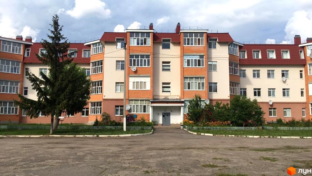 Житловий будинок з червоним дахом, помаранчевими та бежевими секціями фасаду. На прибудинковій території ростуть дерева, кущі та квіти.